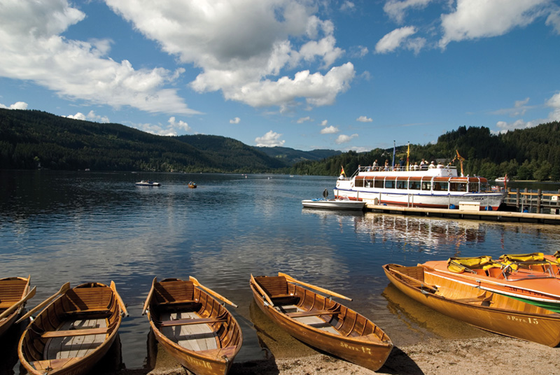 Ein ruhiger See mit mehreren Holzruderbooten, die am Ufer aufgereiht sind. Im Hintergrund liegt ein größeres Boot am Steg, auf dem Menschen zu sehen sind. Um den See herum erstrecken sich grüne Hügel und Wälder, und der Himmel ist teils bewölkt. Die Wasseroberfläche spiegelt die Landschaft und Boote wider.