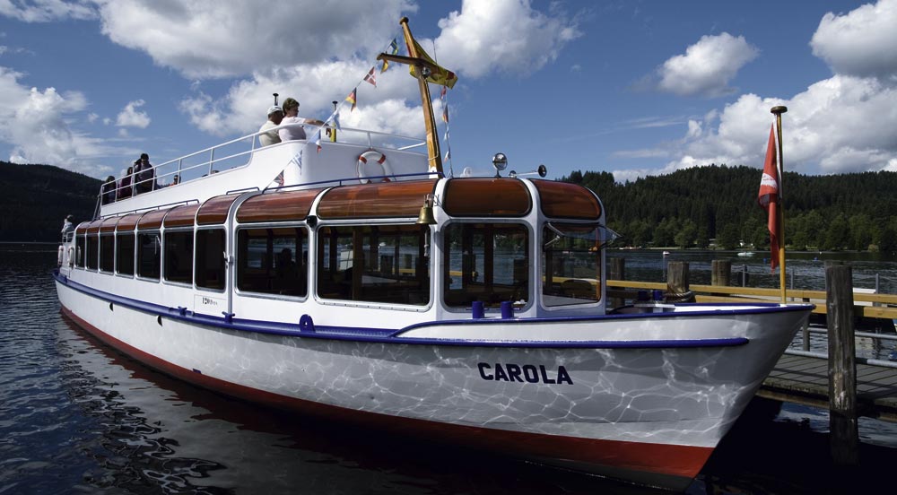 Ein Boot namens ‚CAROLA‘ am Pier eines Sees, mit weißem Rumpf, blauem und rotem Akzent. Menschen stehen auf dem Oberdeck, das mit bunten Flaggen dekoriert ist. Im Hintergrund sind Bäume, Hügel und ein teilweise bewölkter Himmel sichtbar. Das Boot spiegelt sich im ruhigen Wasser.