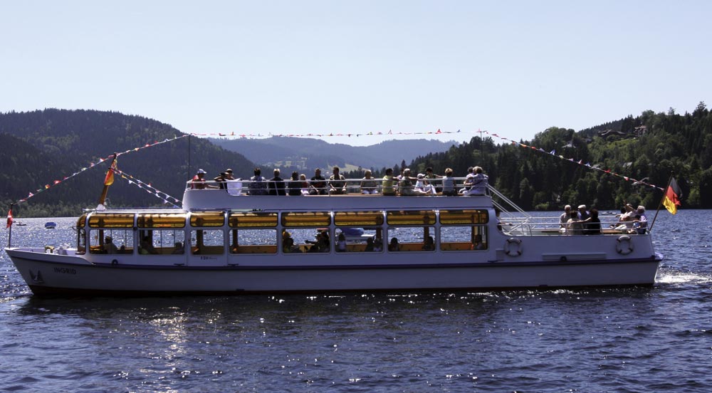 Ein Boot namens ‚Ingrid‘ auf einem See mit zwei Ebenen: die untere ist verglast und geschlossen, die obere offen und mit Sitzplätzen für Passagiere. Das Boot ist mit bunten Flaggen dekoriert. Umgeben von bewaldeten Hügeln und Bergen unter sonnigem Wetter. An den beiden Enden sind deutsche Flaggen befestigt.