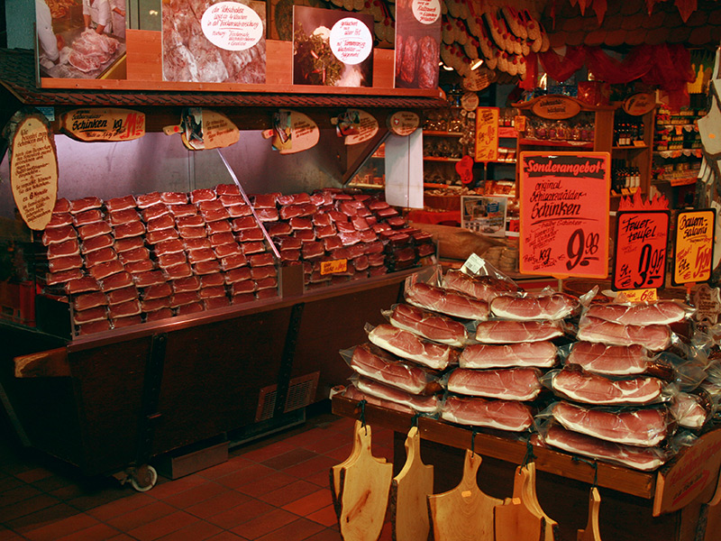 Ein Metzgerei-Bereich mit einer breiten Auswahl an Fleischprodukten. Im Vordergrund sind Tische mit verpackten Fleischsorten wie Schinken und Salami. Dahinter stehen Regale mit weiteren Fleischwaren. Schilder bieten Sonderangebote an, wie z.B. ‚original Schwarzwälder-Schinken‘ für 9,98 Euro und ‚Baum-Salami‘ für 6,95 Euro. Zusätzliche Information zu traditionellen Herstellungsweisen und die Einladung, lokale Spezialitäten zu probieren, ist ebenfalls hervorgehoben.