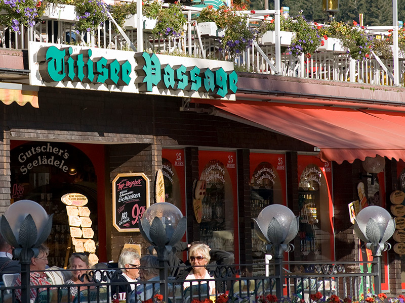 Ein Außenbereich eines Einkaufs- oder Restaurantkomplexes mit dem Schild ‚Titisee Passage‘ in grünen Buchstaben. Im unteren Bereich ist ein Ladeneingang mit dem Namen ‚Gutscher's Seelädle‘ zu sehen, begleitet von einem schwarzen Schild mit weißem Text. Verschiedene Werbeschilder sind präsent, einschließlich eines Angebots für Schinken (100g für 0,99€). Oberhalb des Bereichs sind Blumenkästen mit roten Blumen dekoriert. Menschen sitzen im Außenbereich, umgeben von Straßenlaternen und einem roten Markisendach. Die Szene ist belebt und farbenfroh.