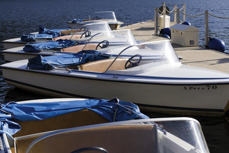 Mehrere kleine Motorboote, die parallel zueinander an einem Pier angedockt sind. Die Boote haben Windschutzscheiben und blaue Abdeckungen über den Sitzbereichen. An der Seite eines Boots steht ‚5 Pers. 70.‘. Das Wasser ist ruhig, und das Pier ist mit Sicherheitsseilen ausgestattet.